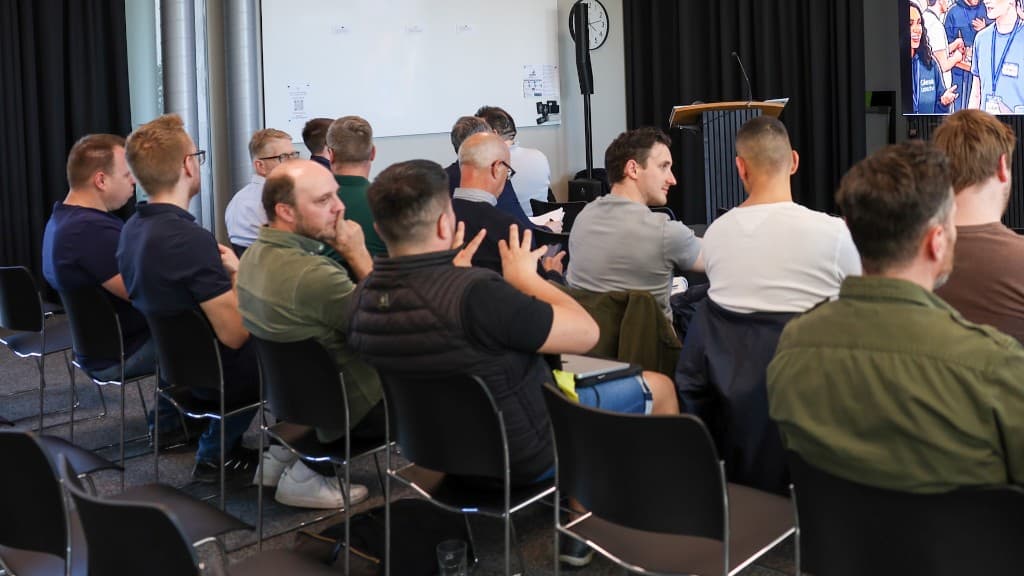 Blick über die Schultern der Zuhörenden Richtung Bühne mit Whiteboard und Bildschirm im GlessHub.