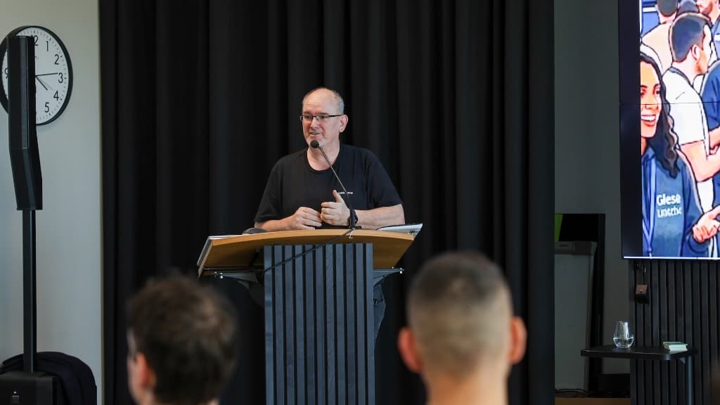 Redner am Pult vor dunklem Vorhang, Präsentation auf dem Seitenmonitor.