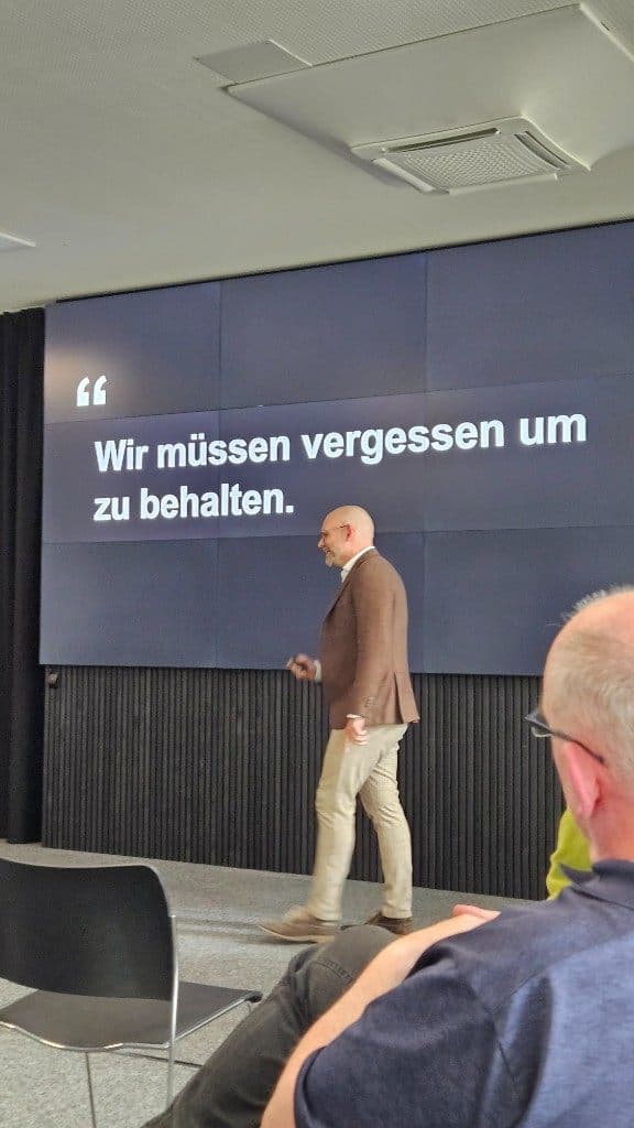 Dominik Volkert geht vor der Leinwand entlang; darauf steht „Wir müssen vergessen um zu behalten.“