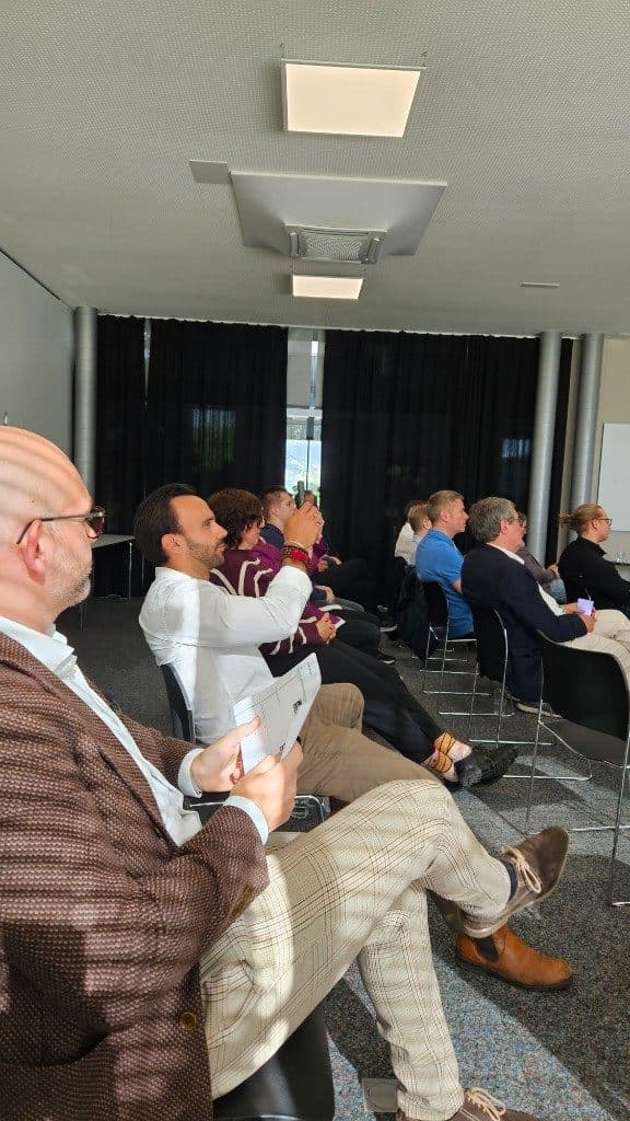Teilnehmende verfolgen den Vortrag im GlessHub; ein Besucher macht ein Foto mit dem Smartphone.